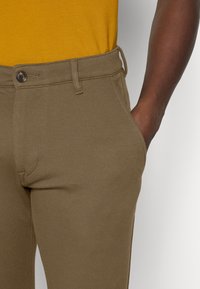 Close-up van een persoon die gestructureerde bruine broek draagt met een knoop en riemlussen, hand rustend in de rechter voorzak.