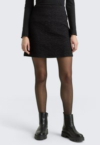 Fekete tweed mini szoknya térbeli anyaggal, karcsú fazonnal és átlapolt kialakítással. Térdzokni fekete bőr csizmával és áttetsző fekete harisnyával párosítva.