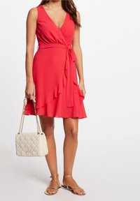 Abito avvolgente rosso con scollatura a V profonda, orlo arricciato e vita con legature. Abbinato a una borsa beige testurizzata e sandali dorati con cinturini.