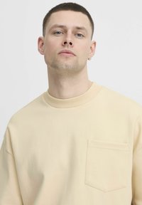 Béžová mikina s okrúhlym výstrihom, rebrovaným golierom a prednou vreckom. Mäkká textúra a voľný strih. Obmedzené pozoruhodné detaily.