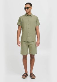 Herre lys grøn kortærmet skjorte med knapper og matchende shorts, lavet af et tekstureret stof, parret med brune sandaler og solbriller.