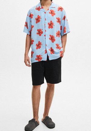 Homme portant une chemise à manches courtes bleu clair à boutons avec des fleurs d'hibiscus rouges, un short noir et des chaussures grises sans lacets.