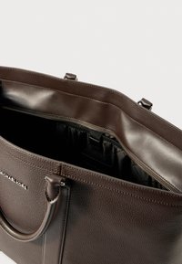 Brązowa skórzana torba tote o teksturowanym wykończeniu, z podwójnymi uchwytami i zasuwaną kieszenią wewnętrzną. Nazwa marki widoczna na boku.