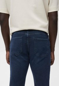 Persona de piel oscura vistiendo una camisa blanca de manga corta y jeans azul oscuro, vista desde atrás con las manos relajadas a los lados.