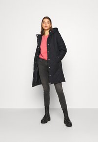 Manteau noir matelassé avec une capuche, doté de boutons pression et de fermetures éclairs sur les côtés, porté sur un haut rose et un jean gris, associé à des bottines noires.