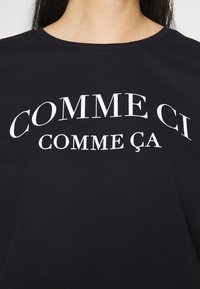 Černé tričko s bílým nápisem "COMME CI" obloukem nad "COMME ÇA" na hrudi, které nosí osoba s dlouhými tmavými vlasy.