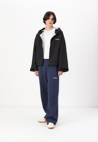Veste noire imperméable avec capuche, doublure grise, poches zippées, associée à un pantalon de survêtement bleu foncé et des baskets beiges. Accents de logo simples.