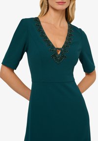 Tyrkysové šaty s krátkými rukávy, výstřihem do V zdobeným černými korálky a detaily ve tvaru klíčové dírky. Hladký materiál s přiléhavou korzetovou částí a rozšířenou sukní.