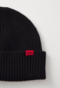 Black ribbed knit beanie featuring a folded cuff and a red rectangular tag with "HUGO" stitched in white. Textured and stretchy material.