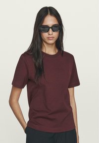 Burgundy cotton t-shirt with short sleeves, crew neckline, smooth texture. Model wearing black sunglasses, standing against a light background.