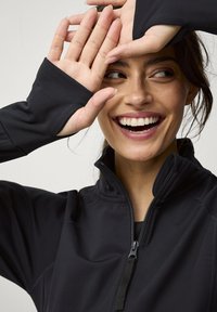 Zwarte, getailleerde zip-up top met een hoge kraag, voorzien van duimgaten in de mouwen en een zachte textuur. Model lacht met handen dicht bij het gezicht.