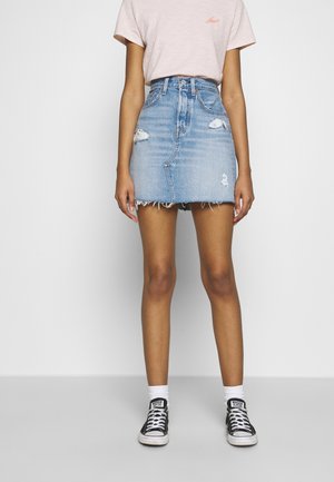 Kvinde iført en lyserød kortærmet skjorte, en distressed blå denim mininederdel, hvide sokker og sorte Converse-sneakers, stående op ad en hvid væg.