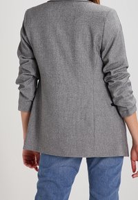 Szary marynarka o teksturowanej fakturze z dopasowanym krojem, pokazana z tyłu. Ma podwinięte rękawy i jeden rozcięcie u dołu. Nosi się ją na jasnoniebieskie dżinsy.