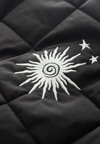 Fekete steppelt szövet, középen egy fehér hímzett napmintával, forgó sugarakkal, kiegészítve kis csillagmotívumokkal.