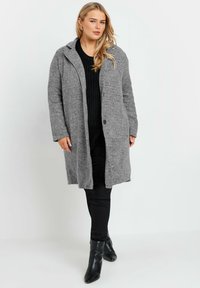 Grå lång kappa i ett mjukt, texturerat tyg med knappstängning, sidofickor och huva, tillsammans med en svart tröja och byxor.