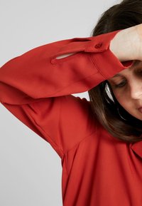 Blouse rouge à manches longues, en tissu lisse. Comprend une fermeture à bouton et un détail découpé  à la manche. Design simple.