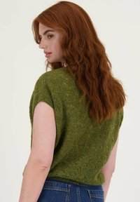 Groene gebreide top met korte mouwen, voorzien van een textuur en een patroon. Zichtbare stiksels en een losse pasvorm, gecombineerd met blauwe spijkerbroek.