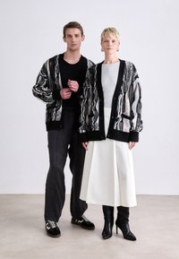Two models stand side by side. Each wears a patterned, oversized black and white cardigan. One model has dark trousers, the other a white skirt.