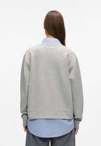 Άτομο με μακριά καστανά μαλλιά φοράει γκρι φούτερ πάνω από μπλε καρό πουκάμισο, στραμμένο μακριά σε έναν λευκό ομοιόμορφο φόντο.