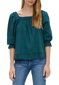 Kobieta w tealowej, fakturowanej bluzce z kwadratowym dekoltem i trzyczwartymi bufiastymi rękawami, zestawionej z niebieskimi dżinsami.