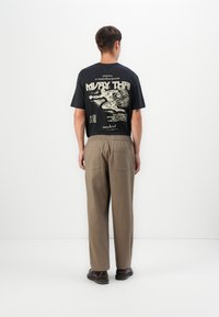 Mann steht von hinten und trägt ein schwarzes Muay Thai T-Shirt mit Grafik, beigefarbene Hose und dunkelbraune Schuhe vor einem schlichten weißen Hintergrund.