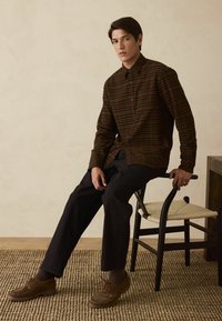 Camicia marrone a quadri con bottoni, pantaloni scuri e scarpe in camoscio marrone. Uomo seduto su una sedia nera con seduta intrecciata, sfondo neutro e texture.