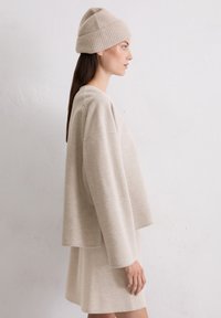 Beżowy wełniany sweter w oversize z długimi rękawami, nałożony na dopasowaną spódnicę. Połączony z ryflowaną beżową czapką. Gładka tekstura.