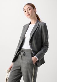Grijze geruite blazer met twee knopen, gecombineerd met grijze broek met een elastische tailleband en zijstreep. Gekocht over een wit top.