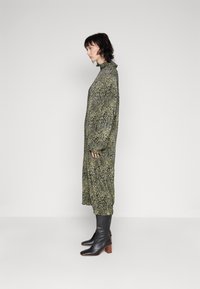 Robe longue et ample de couleur verte avec un imprimé léopard noir, en tissu texturé, col montant et manches longues ; assortie à des bottes à talons noires.