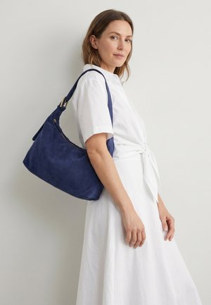 Femme en robe blanche portant un grand sac à épaule bleu marine, debout devant un fond uni gris clair.
