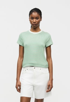 Γυναίκα που φοράει κοντομάνικη μπλούζα με πράσινες και άσπρες ρίγες και λευκά τζιν σορτς, στέκεται μπροστά σε απλό φωτεινό φόντο.