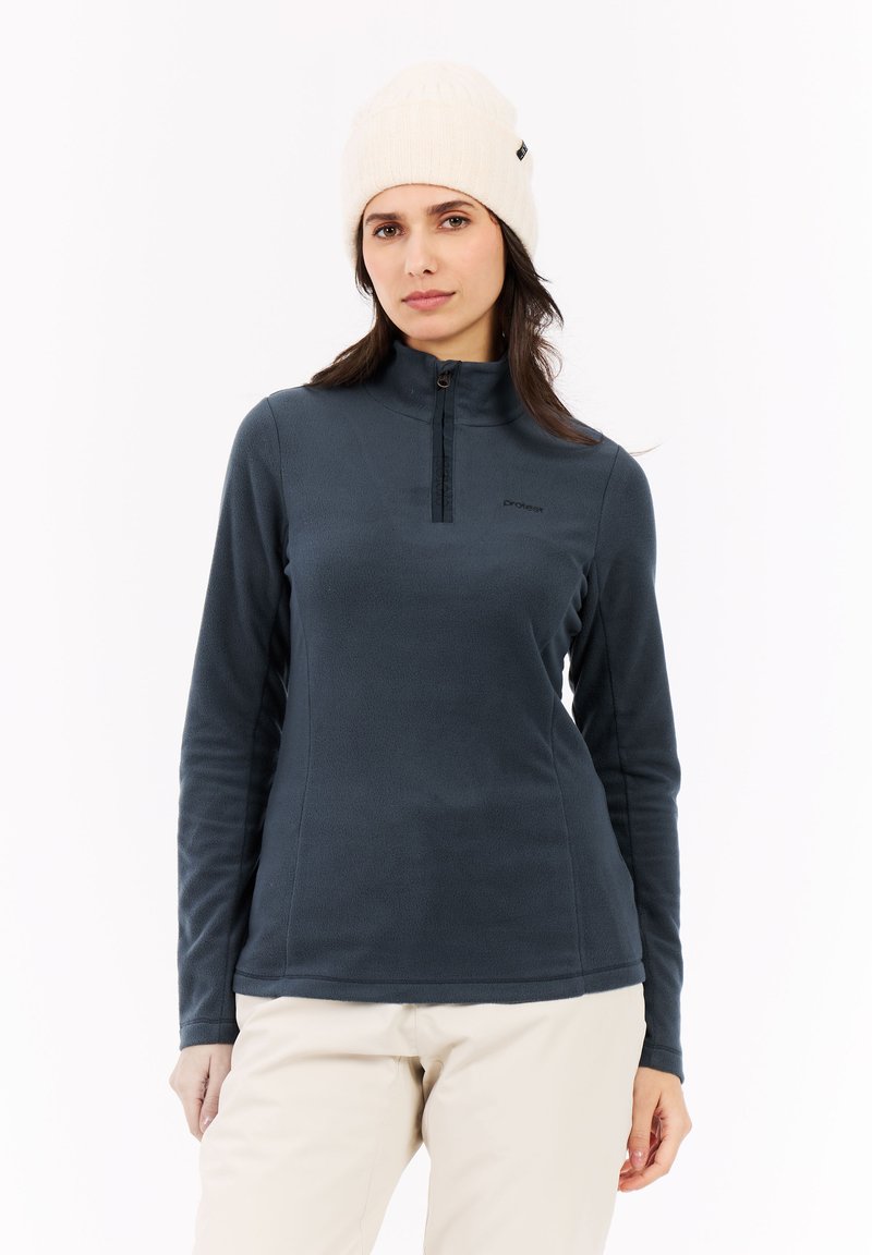 Donna con berretto di maglia bianco, pullover in fleece blu scuro con zip a quarti, e pantaloni beige su uno sfondo bianco, in piedi e rivolti in avanti.