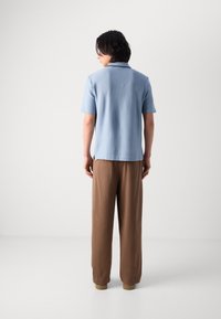 Polohemd aus leichtblauem Strickstoff mit kurzen Ärmeln; kombiniert mit locker sitzenden braunen Hosen. Verfügt über einen klassischen Kragen und seitliche Schlitze.