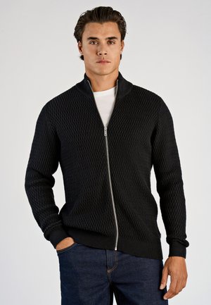 Mand iført en sort tekstureret lynlås-sweater over en hvid skjorte med mørkeblå jeans, stående mod en ensartet lys baggrund.