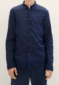 Mörkblå långärmad skjorta med krage, som har knäppning fram, manschettdetaljer och en slät tygtextur.