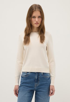 Giovane donna con lunghi capelli castani indossa un maglione crema a maniche lunghe e jeans blu, in piedi davanti a uno sfondo chiaro e semplice.