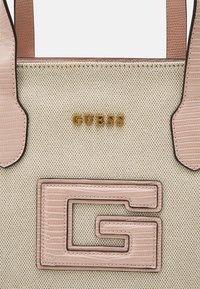 Beżowa torba na ramię w wzory z różowymi, teksturowanymi uchwytami. Złote logo "GUESS" oraz duży różowy akcent w kształcie litery "G" umieszczone na przodzie.