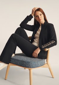 Chaqueta negra con cremallera y detalles de cristal en la manga, combinada con pantalones negros. El modelo se sienta en una silla de madera con un cojín azul texturizado.