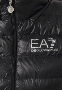 Czarna ocieplana kurtka o błyszczącej powierzchni, z zamkiem błyskawicznym z przodu oraz srebrnymi logotypami EA7 i Emporio Armani na klatce piersiowej.