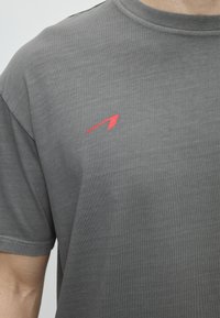 Κοντινό πλάνο ενός ατόμου που φοράει γκρι μπλουζάκι με στρογγυλή λαιμόκοψη και μικρό κόκκινο γωνιακό λογότυπο στο αριστερό στήθος.