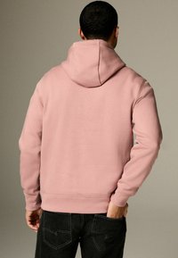 Sudadera rosa con un bolsillo canguro, puños y dobladillo de canalé, y capucha con cordón. La tela parece suave y texturizada. Se llevan jeans negros.