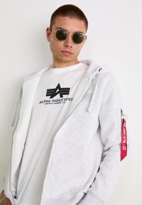Šedá mikina na zips s prednou kapsou a červeným textilným štítkom, oblečená cez biele tričko s čiernym logom, doplnená okrúhlymi slnečnými okuliarmi.