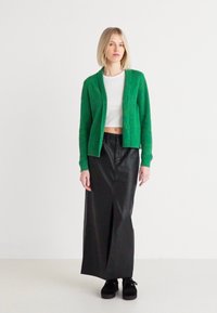 Cardigan vert à côtes, style court, porté sur un top blanc. Associé à une jupe en cuir noire avec une fente devant. Chaussures à plateforme noires.