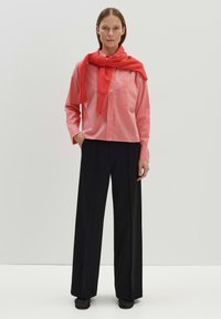 Chemise boutonner rose clair superposée à un pull rouge noué autour du cou, accompagnée d'un pantalon large noir et de chaussures noires.