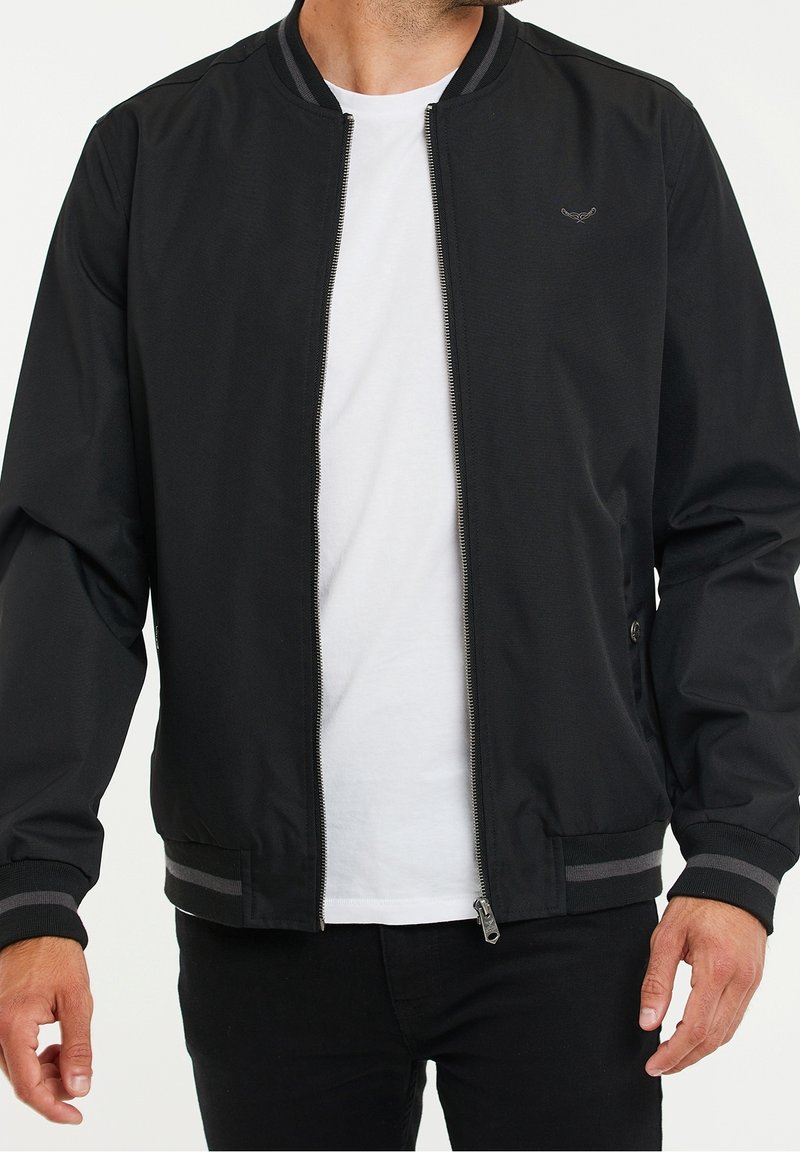 Homme portant une veste bomber noire avec des poignets rayés gris et un t-shirt blanc, mains détendues sur les côtés, torse visible, tête coupée.