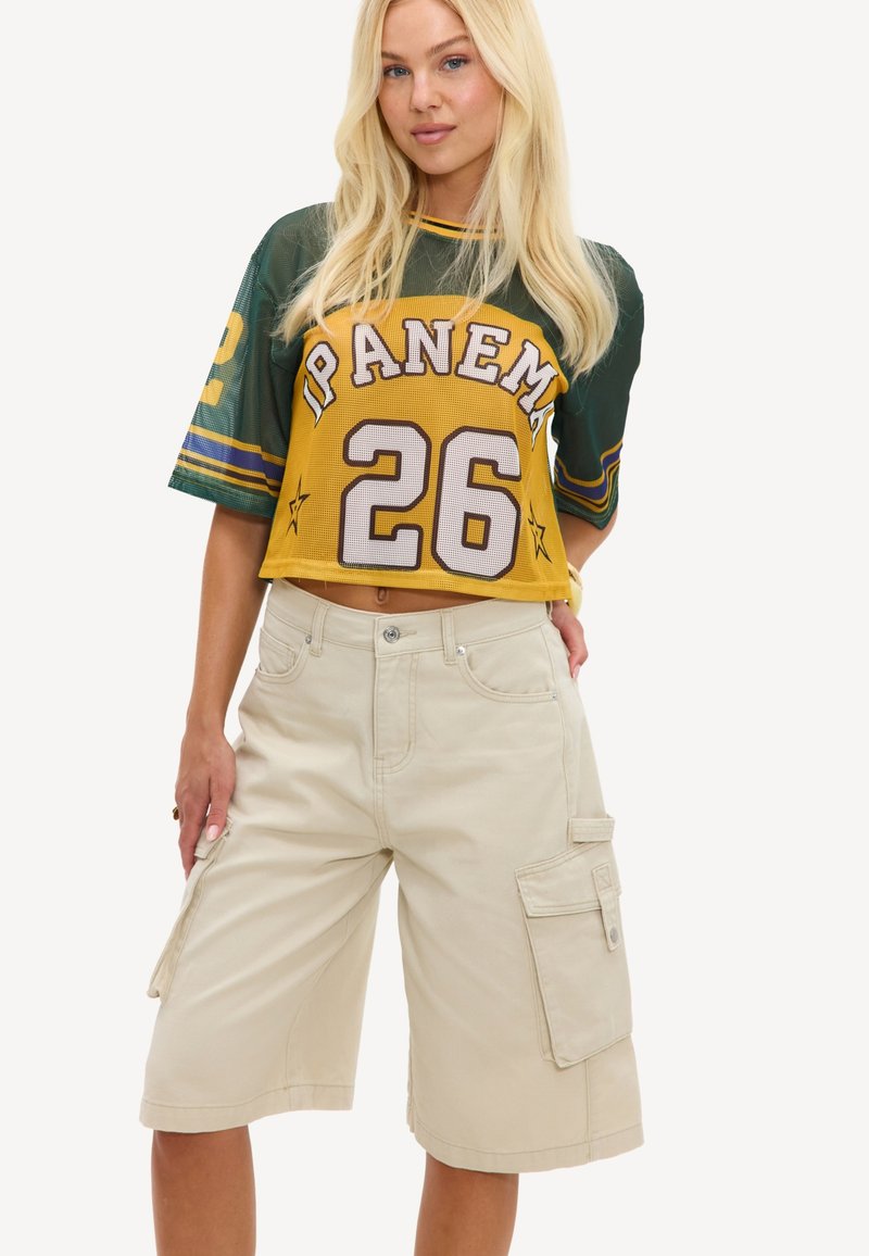 Jeune femme portant un crop top vert et jaune avec "Ipanema 26" et un short cargo beige longueur genou, debout, une main dans la poche.
