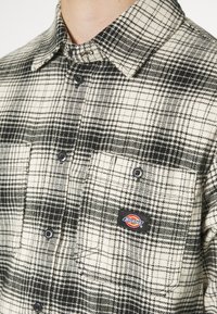 Chemise à carreaux noire et blanche à boutons, avec poches poitrine, dont une portant un patch logo de la marque Dickies.