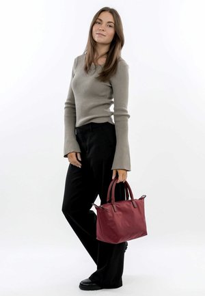 Sac à main bordeaux avec des accents en cuir, de forme structurée et deux poignées courtes. Le modèle porte un haut beige à côtes et un pantalon noir.