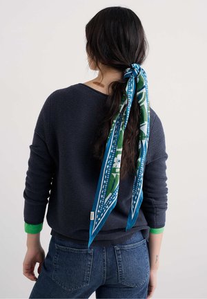 Blauw-groen patroon sjaal in een paardenstaart, gedragen met een marineblauwe trui en spijkerbroek. De trui heeft groene accent manchetten.