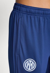 Mørkeblå treningsbukser med elastisk midje, sidelommer og et brodert rundt logo på låret for enkel identifikasjon.